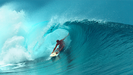 CLOSE UP: Young professional surfboarder finishes riding another epic tube wave on a sunny day in French Polynesia. Surfer having fun in the refreshing emerald water on a perfect day for surfing.