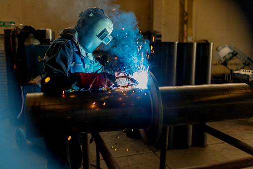 A man in personal protective equipment wearing a welding helmet is using a machine to weld a piece of metal in an engineering factory