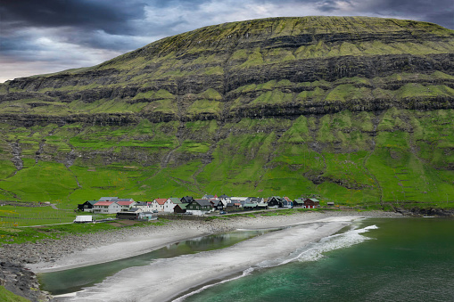 AERIAL: Dark clouds gather over the serene oceanfront village in the picturesque green valley. Stunning background of a tranquil town protected by steep hill in the scenic countryside of Faroe Islands