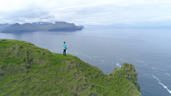 AERIAL, COPY SPACE: Flying over beautiful Scandinavian landscape and unrecognizable female photographer observing the vast sea. Spectacular aerial shot of the rugged seaside in scenic Faroe Islands.
