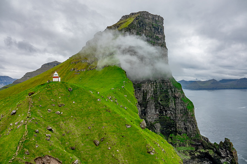 AERIAL: Flying towards a small white lighthouse on top of the grassy hill in Faroe Islands. White clouds drift along the rocky cliffs on a picturesque fjord in the scenic Scandinavian wilderness.