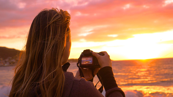 COPY SPACE, CLOSE UP: Unknown female photographer with wind in her hair filming the orange sunset from a scenic beach. Cinematic shot of woman taking photos of the ocean on a stunning summer evening