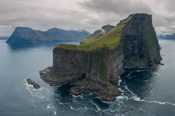 istock AERIAL: Flying around a grassy islet with black rocky cliffs towering over sea. 1182236286