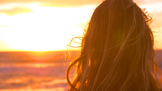 LENS FLARE, COPY SPACE, CLOSE UP: Unrecognizable young female photographer looking at the beautiful evening sun and the small sea waves splashing towards the beach. Cinematic view of girl and sunset.