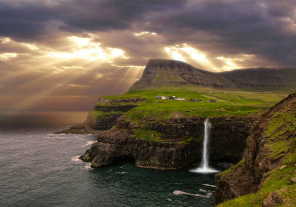 AERIAL: Bright golden sun rays shine through the dense clouds above the breathtaking waterfall in rural part of Faroe Islands. Spectacular shot of sun shining on vibrant green coastal countryside.