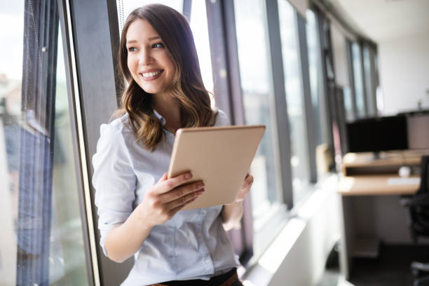 窓の前に立ちながらデジタルタブレットを使った魅力的なビジネスウーマン - business office business person window ストックフォトと画像