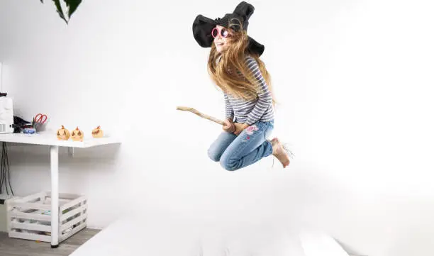 Photo of Girl in a witch hat jumps on a mattress with a broom.