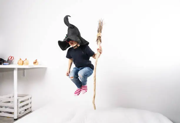 Photo of Boy in a witch hat jumps on a mattress with a broom.