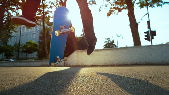 LOW ANGLE, LENS FLARE, CLOSE UP: Unknown skateboarder jumps and lands a cool trick while riding on concrete sidewalk in sunny city. Cinematic view of skateboard flipping in air underneath man's feet.