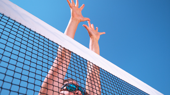 LOW ANGLE, CLOSE UP: Young female beach volleyball player jumps up to block the ball coming towards her. Fit Caucasian woman playing volleyball during her summer vacation jumps high up in the air.