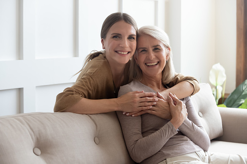 Loving adult 30s daughter hug elderly mother from behind while mom sitting on couch people posing looking at camera smiling feels happy, concept of multi generational family, relative devoted person