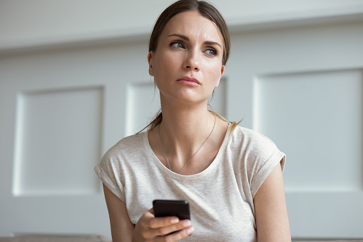 Pensive thoughtful young 30s woman holding smartphone look away lost on sad thoughts waiting first step from man, call or text message or date invitation from boyfriend feels jealousy and concerned