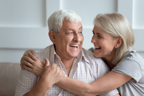Elderly hoary couple embracing sitting on couch indoors Cheerful 80s elderly husband and attractive blond 60s wife sitting on couch laughing embracing indoors, different generations family middle-aged female cuddling her senior father showing love concept father daughter 80s stock pictures, royalty-free photos & images