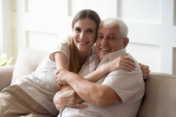 Adult granddaughter and elderly grandfather embracing sitting on couch indoors Adult granddaughter and elderly 80s grandfather sitting together on couch posing look at camera, loving intergenerational family portrait warm love and care between people different generation concept father daughter 80s stock pictures, royalty-free photos & images