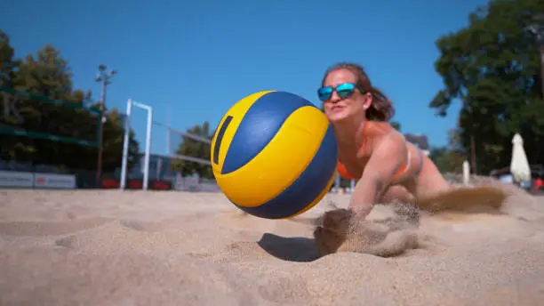 CLOSE UP, DOF: Athletic Caucasian girl playing beach volleyball jumps into the sand and strikes the ball with her hand. Cinematic shot of a fit young female volleyball player saving a game point.