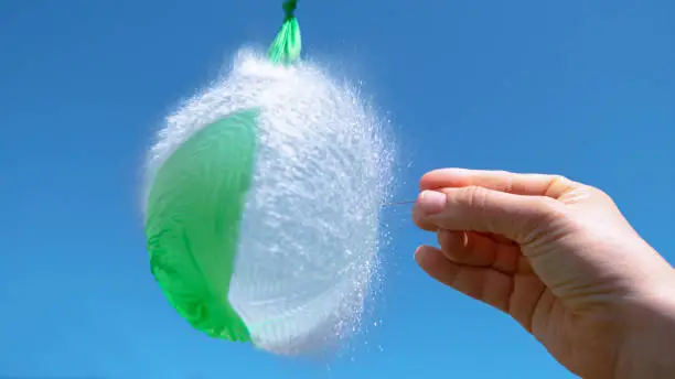 CLOSE UP: Unrecognizable person holding a needle pierces a green balloon full of water. Hand holding a metal pin bursts a rubber balloon filled with a crystal clear liquid. Water droplets flying.