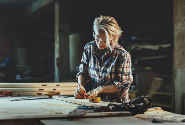 Attractive middle aged woman carpenter designer works with ruler, make notches on the tree in workshop.  Image of modern femininity. Concept of professionally motivated women stock photo