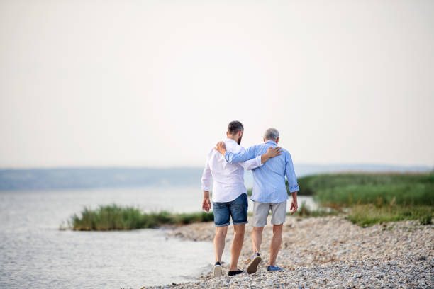 rear view of senior father and mature son walking by the lake. copy space. - men beach back rear view imagens e fotografias de stock