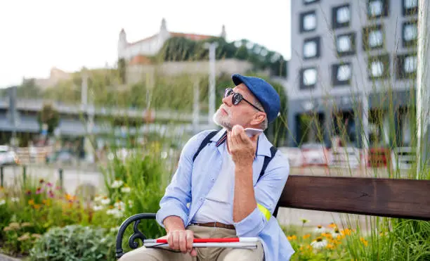 A senior blind man with smartphone sitting on bench in park in city, making phone call.