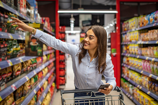 Photo of of young cheerful woman in supermarket with shopping trolley choosing products and using phone. Looking aside. The housewife chooses food for dinner at home on the background of shelves in a supermarket.