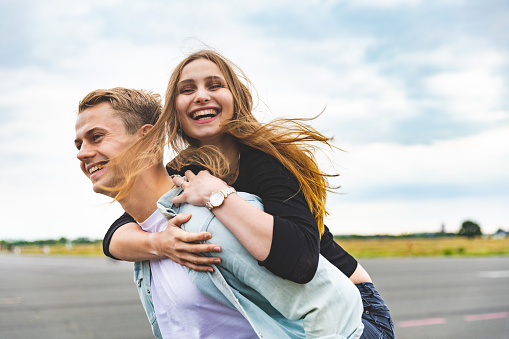 Happy couple having fun and enjoying a piggyback ride - Caucasian man and woman, best friends, sharing time together and laughing in Berlin at Tempelhof airport park - Friendship and happiness lifestyle concepts