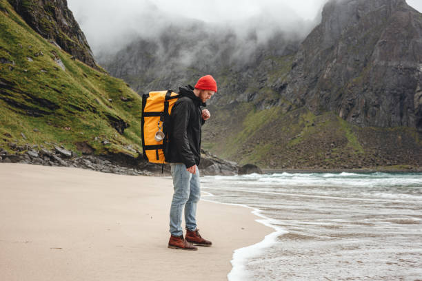 man adventurer walking by the sea beach among cloudy mountains. traveling alone by the ocean sand heathy lifestyle active vacations - rock norway courage mountain imagens e fotografias de stock