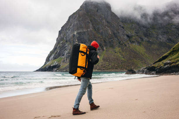 turista sozinho que anda ao longo da praia do mar entre montanhas nevoentas. viajante da aventura com a trouxa que caminha pela areia do oceano - rock norway courage mountain - fotografias e filmes do acervo