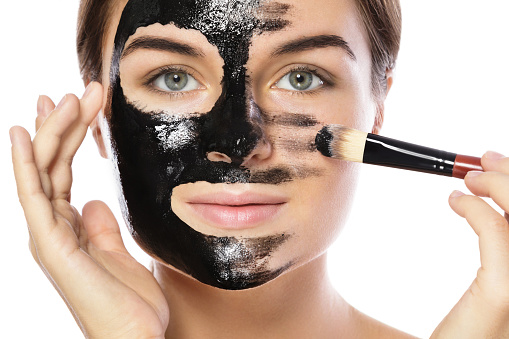 Beautiful woman is applying purifying black mask on her face over white background