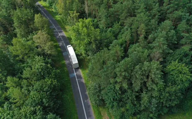 Photo of Aerial drone perspective view on white truck with cargo trailer riding through the forest on curved asphalt road