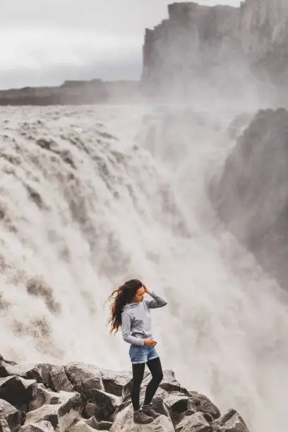 Photo of Young woman in jeans shorts and gray hoodie with dramatic view of famous Iceland waterfall Dettifoss. Breathtaking landscape, stream of water, most powerful waterfall in Europe. Small person, big waterfall.