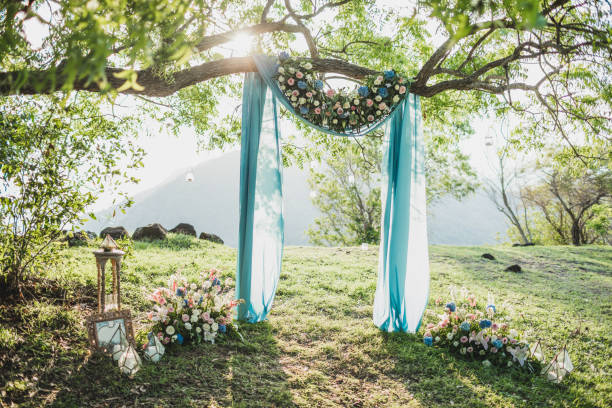 istock Sunset wedding ceremony arch with flower decoration and blue cloth canopy hanging on big tree. Blue hydrangea, pink roses, lily. Pastel colored. 1182382164