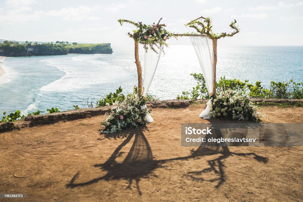 Arco de ceremonia de boda al atardecer con ramas de madera y decoración floral y dosel de tela blanca. Vista del acantilado de Balangan en Bali. Boda con vista al mar. - Foto de stock de Acantilado libre de derechos