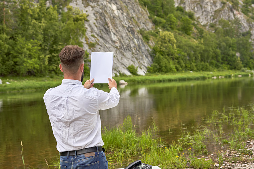 A man in a white shirt, tie and Santa hat holds in his hand a blank sheet of paper on the background of a mountain river on a summer day.