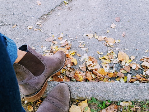 Ankle boots chelsea on legs in jeans on a background of asphalt and yellow autumn leaves.