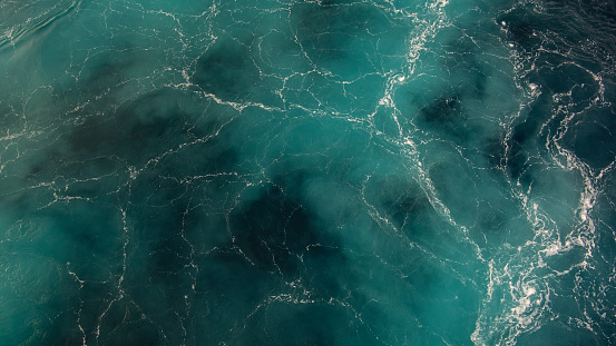 Close-up view of turquoise blue sea or ocean waves creating a marble effect with white flowing lines and blue marine, navy tones visible in the crystal clear water, natural background with copy space.