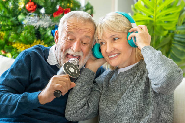 homme et femme aînés de couples chantant la chanson de noel ensemble, s'asseyant sur le sofa avec l'arbre et la décoration à l'arrière-plan - christmas tree audio photos et images de collection