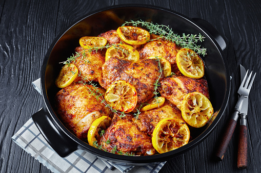 baked Chicken Thighs With Roasted Lemon Slices and thyme in a black ceramic dutch oven on a wooden table, horizontal view from above