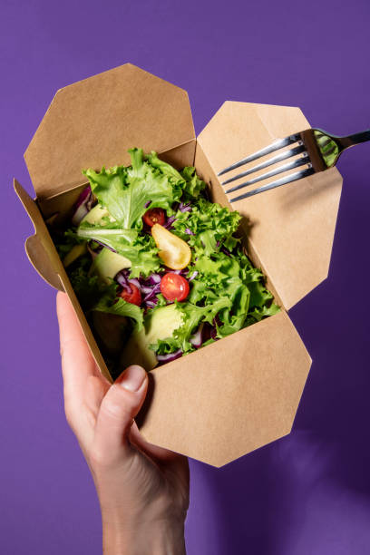 istock Woman's hand holds a mixed salad in cardbox with fork other hand. 1182402580