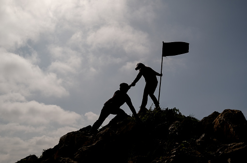 Silhouette image of one friend with flag try to pull his other friend over the cliff of mountain and they climb and help each other with concept teamwork for successful.