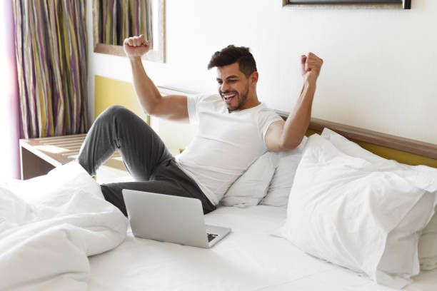 istock Happy man raising hands up, sitting on bed, using laptop 1182415409
