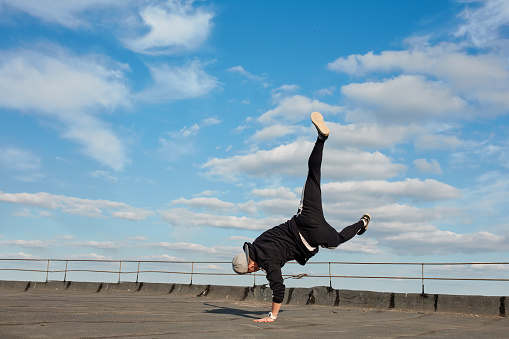Street dancer hipster guy makes back flip with arm support. Asian Breakdancer exercising on roof. Low angle mid-air view against blue sky background