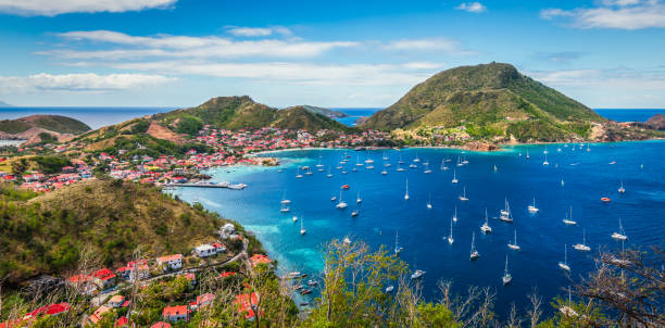 Panoramic landscape view of Terre-de-Haut Island, Guadeloupe, Les Saintes. Colorful image of Guadeloupe Terre de Haut bay and town with buildings along the coastline. Small ships anchored in port. Cruise destination. Green mountains in the background. Blue sky and some white clouds. caribbean islands stock pictures, royalty-free photos & images