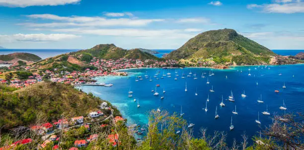 Colorful image of Guadeloupe Terre de Haut bay and town with buildings along the coastline. Small ships anchored in port. Cruise destination. Green mountains in the background. Blue sky and some white clouds.