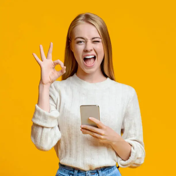 Photo of Millennial girl holding smartphone, winking and gesturing ok