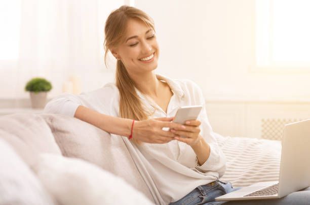 istock Relaxed Woman Using Smartphone Sitting With Laptop On Couch Indoor 1182427385