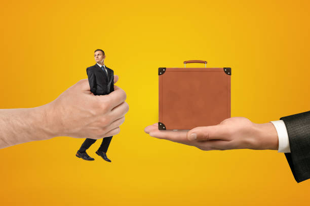 istock Man's hand exchanging tiny businessman for tiny brown travel case held in another man's hand on amber background. 1182430351