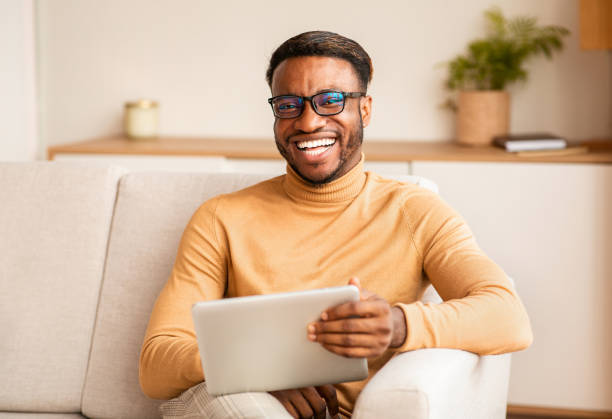 istock Afro Man Holding Digital Tablet Sitting On Couch At Home 1182432142