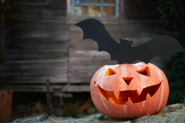 istock Halloween pumpkin head lantern with eyes and a face on a stump, in a rustic setting with a bat on a background of a wall of wooden planks 1182432556