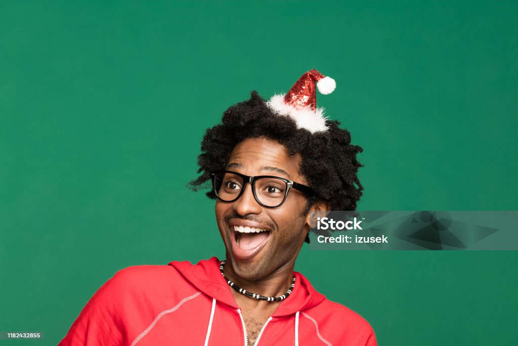 Funny christmas portrait of excited young man wearing in red Funny Christmas portrait of young afro American man wearing Santa Claus hat headband and red blouse, looking away and laughing. Studio shot against green background. Cool Attitude Stock Photo