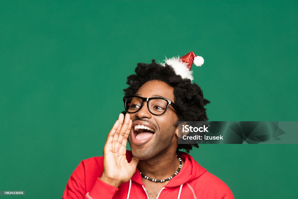 Funny christmas portrait of excited young man wearing in red Funny Christmas portrait of young afro American man wearing Santa Claus hat headband and red blouse, looking away and shouting. Studio shot against green background. 25-29 Years Stock Photo